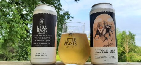 A glass of beer in between a black can of beer and a brown can of beer. All three are siting on a wood table outside with trees in the background. The glass as well as both bottles have the text LITTLE BEASTS on them.